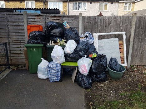 A collection of black, white, and patterned rubbish bags filled with household waste is piled on a small paved area adjacent to a wooden fence in an outdoor setting. The bags are stacked haphazardly, with some leaning against a green wheelie bin and others resting directly on the ground. Behind the rubbish, a white-framed object that appears to be a dismantled bed frame or similar furniture component is partially visible, leaning against the fence. To the right, there is a round, green plastic container without a lid, and on the left, an orange waste bin is positioned next to the wooden fencing. The background shows the upper floors of residential buildings, indicating a suburban or urban environment. The scene suggests a typical rubbish collection or clearance area, relevant to services provided by Office Clearance Gipsy Hill in the local postcode area.