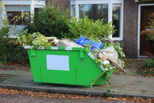 A large green skip positioned on a paved driveway in front of a brick residential building with white-framed windows, filled with various types of rubbish including tree branches, leafy green foliage, cardboard boxes, plastic bags, and miscellaneous waste materials. The skip is partially loaded, with some waste spilling over its edges, and is situated beside a curb with fallen leaves scattered on the pavement. Surrounding the property are bushes and shrubs, indicating a suburban or residential area typical of Gipsy Hill. This scene illustrates a waste collection or rubbish removal service, such as that provided by Office Clearance Gipsy Hill, in a local neighbourhood context, emphasizing the handling of garden and household waste for clearance. The environment appears natural daylight with clear visibility of texture and color details in the waste and surrounding greenery.