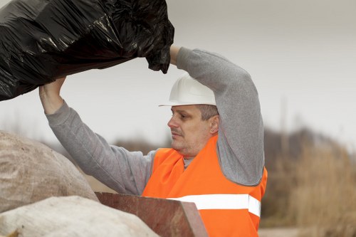 A man wearing a white safety helmet and an orange high-visibility vest is seen outdoors on a cloudy day, lifting a large black rubbish bag above his head. He appears focused and is standing in a rural or semi-industrial setting, with blurred natural background behind him. The black bag is made of heavy-duty plastic, filled with waste, and partially obscures his face as he manages the removal. Next to him, there are other rubbish removal materials, possibly including conjoined bags or bundled waste. The scene suggests a rubbish collection or clearance operation, aligning with services offered by Office Clearance Gipsy Hill for waste disposal and clearance tasks. The overcast sky and open environment provide natural, diffuse lighting, emphasizing the textures of the waste and safety gear. This image exemplifies professional rubbish handling in a typical outdoor setting within the Gipsy Hill area, highlighting the manual effort involved in waste clearance.