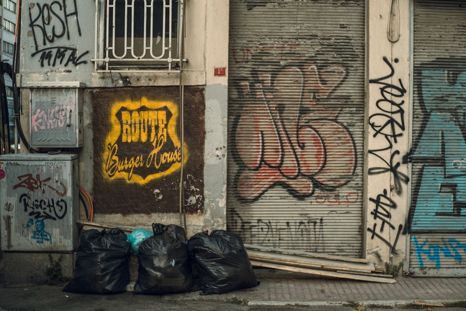 In front of a red-brick building housing a bar and restaurant, there is a black wheeled waste container with a lid, marked with white letters indicating it is for commercial waste collection. The cont