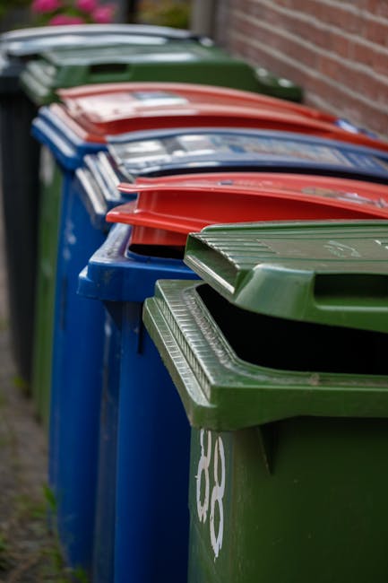 A tall, rectangular blue plastic rubbish bin with a closed lid, positioned outdoors on a patch of grass near a paved area, with a yellow and blue sticker reading 'Fight Back!' affixed to its front. To