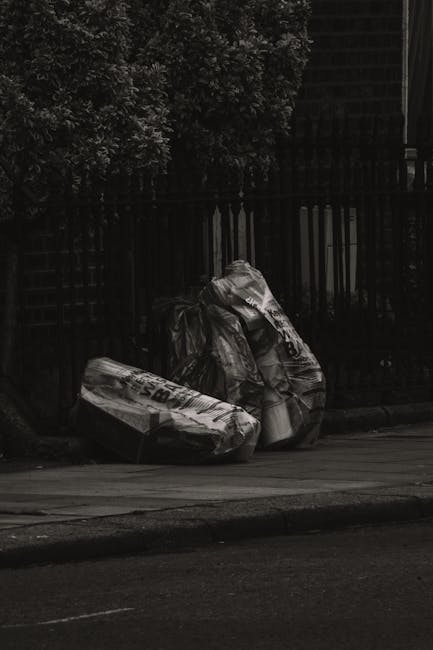 Three large black rubbish bags made of plastic or similar material, filled and somewhat lumpy in appearance, are positioned along the edge of a paved sidewalk, adjacent to a low curb. The bags are pla